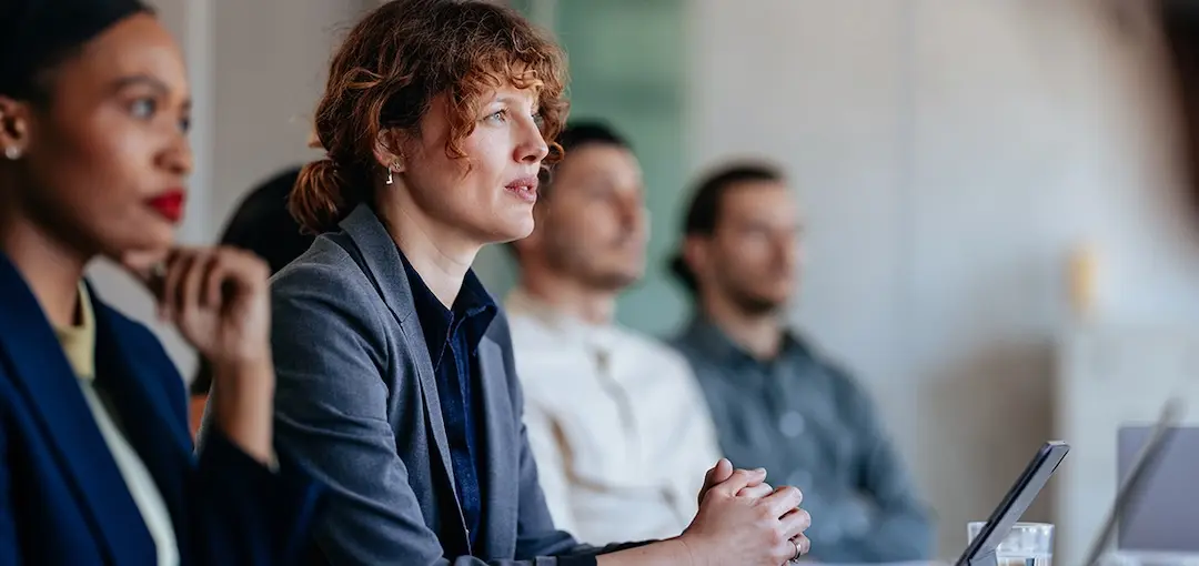 A woman sits at a conference table between another woman and two men.