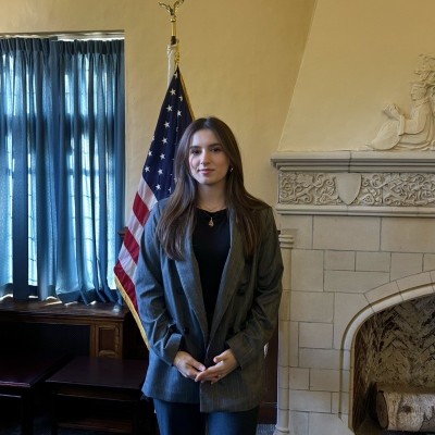 Young woman with long dark hair standing in a room with a U.S. flag behind her.