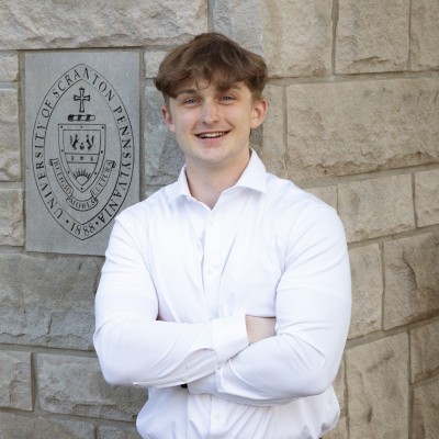 Young man with brown hair in white dress shirt, stands in front of stone wall. 